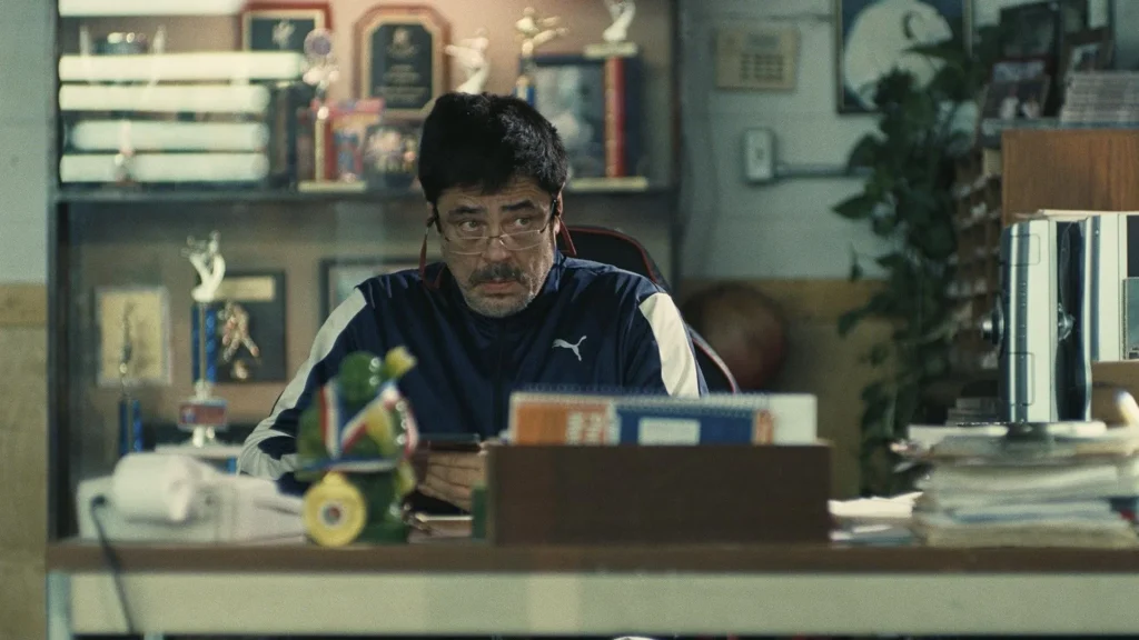 Benicio del Toro em cena de 'Uma Batalha Após a Outra', sentado em uma mesa de escritório com troféus ao fundo, usando uma jaqueta esportiva e óculos, olhando para o celular com expressão séria.