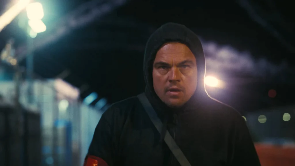 Leonardo DiCaprio em cena de 'Uma Batalha Após a Outra', com expressão intensa, usando capô e casaco, caminhando em um ambiente urbano à noite com luzes dramáticas ao fundo.