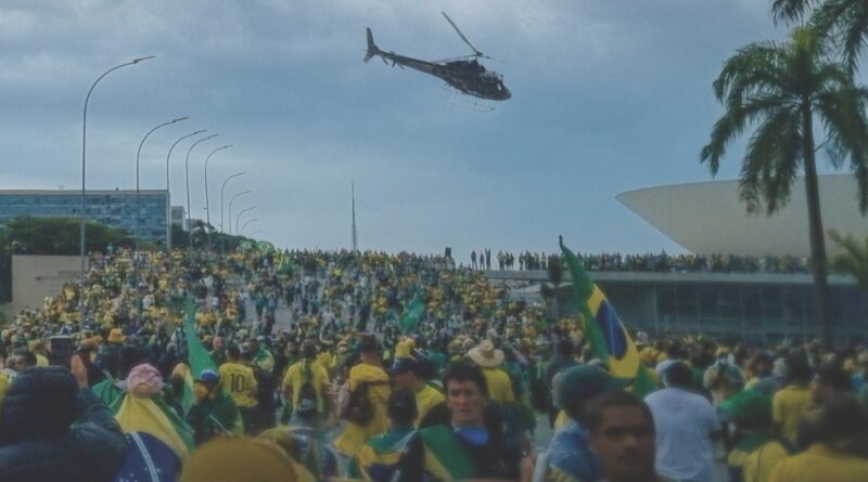 Pôster do filme No Céu da Pátria Nesse Instante, mostrando uma manifestação em Brasília com milhares de pessoas vestindo roupas amarelas e verdes, segurando bandeiras do Brasil, enquanto um helicóptero sobrevoa o local em frente a um prédio icônico. Imagem retirada do nosso blog Multi Nerdz.