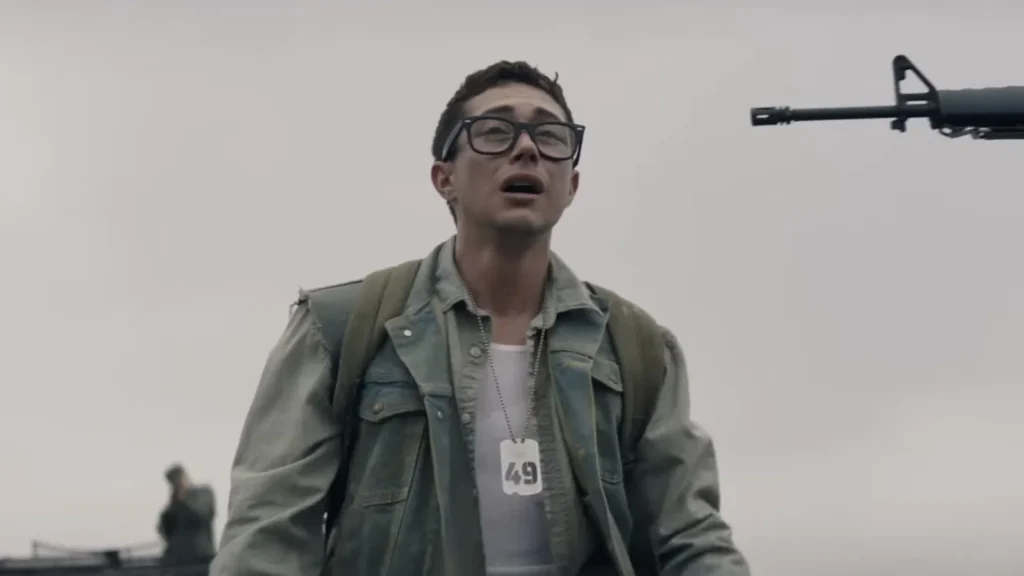 Personagem do filme A Longa Marcha: Caminhe ou Morra caminha exausto enquanto é vigiado por uma arma apontada.