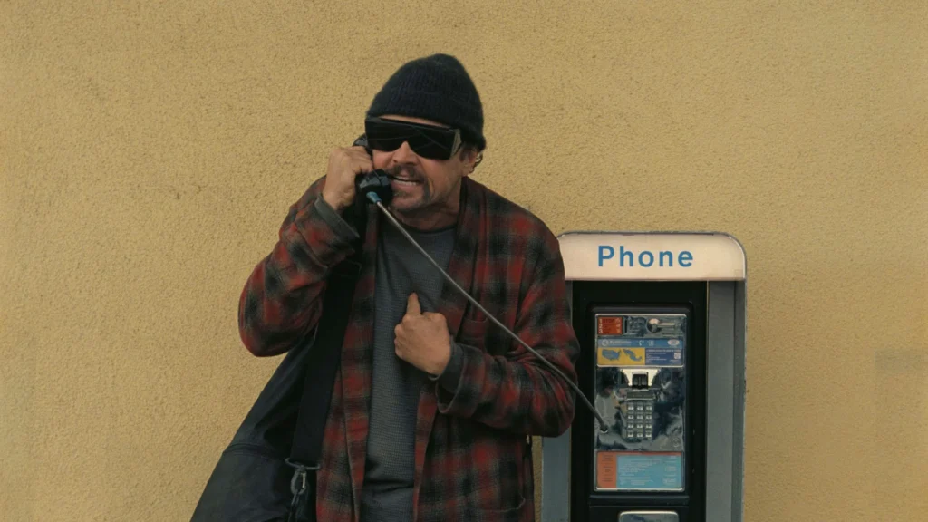 Leonardo DiCaprio em cena de 'Uma Batalha Após a Outra', falando ao telefone público com expressão de raiva, usando uma camisa xadrez e óculos escuros, em frente a uma parede amarela.