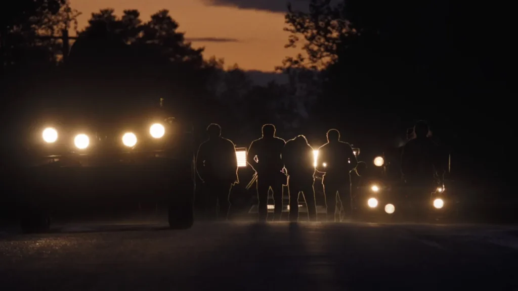 Competidores marcham à noite iluminados pelos faróis dos veículos em cena do filme A Longa Marcha: Caminhe ou Morra.
