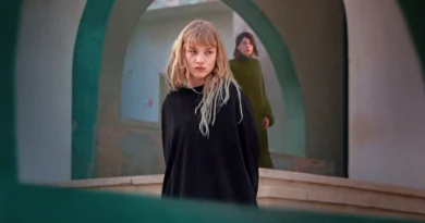 Pôster do filme Rabia: As Esposas do Estado Islâmico, mostrando uma mulher com cabelo loiro e roupas escuras, em primeiro plano, enquanto outra mulher aparece ao fundo, vestindo verde. A imagem captura uma atmosfera de tensão e introspecção, com arcos arquitetônicos ao redor. Crítica de Rabia no canal Multi Nerdz.