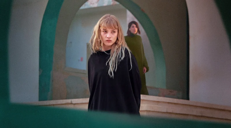 Pôster do filme Rabia: As Esposas do Estado Islâmico, mostrando uma mulher com cabelo loiro e roupas escuras, em primeiro plano, enquanto outra mulher aparece ao fundo, vestindo verde. A imagem captura uma atmosfera de tensão e introspecção, com arcos arquitetônicos ao redor. Crítica de Rabia no canal Multi Nerdz.