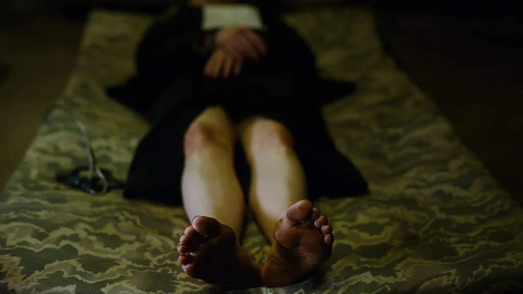 Pés de Emma Stone (Emma Stone) estendidos sobre uma cama de tecido escuro em cena do filme Bugonia, com iluminação sombria e atmosfera tensa. Conteúdo do Multi Nerdz.