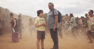 Sergi López e Brúno Nuñez em cena do filme Sirāt em um cenário desértico com poeira levantada. O ator mais velho usa camiseta cinza e mochila, enquanto a criança veste camiseta com estampa de motocicleta, cercados por outras pessoas em um ambiente árido.