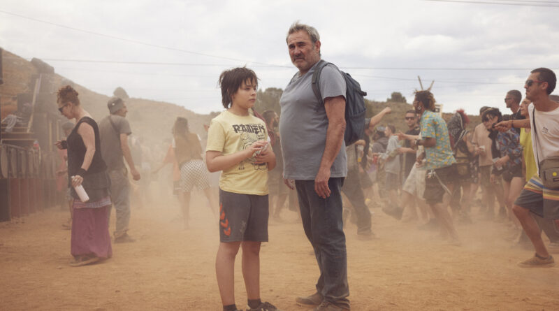 Sergi López e Brúno Nuñez em cena do filme Sirāt em um cenário desértico com poeira levantada. O ator mais velho usa camiseta cinza e mochila, enquanto a criança veste camiseta com estampa de motocicleta, cercados por outras pessoas em um ambiente árido.