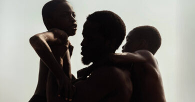 A capa oficial do filme A Sombra do Meu Pai, mostra os atores Ṣọpẹ́ Dìrísù, Chibuike Marvellous Egbo e Godwin Egbo em um abraço coletivo na praia. Eles estão em silhueta, com os corpos molhados brilhando contra um fundo branco e estourado, o que destaca o contorno de seus rostos e o gesto de afeto. A fotografia em preto e branco de alto contraste cria um clima poético e atemporal.