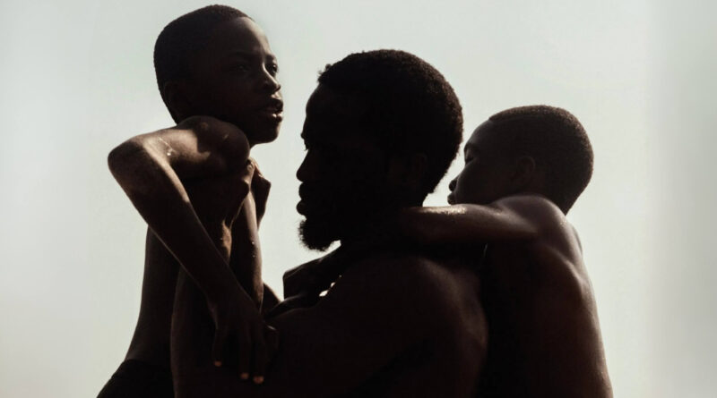 A capa oficial do filme A Sombra do Meu Pai, mostra os atores Ṣọpẹ́ Dìrísù, Chibuike Marvellous Egbo e Godwin Egbo em um abraço coletivo na praia. Eles estão em silhueta, com os corpos molhados brilhando contra um fundo branco e estourado, o que destaca o contorno de seus rostos e o gesto de afeto. A fotografia em preto e branco de alto contraste cria um clima poético e atemporal.