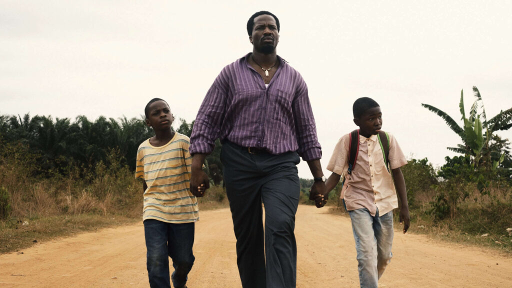 Cena de A Sombra do Meu Pai mostrando Ṣọpẹ́ Dìrísù como o pai caminhando de mãos dadas com os filhos, interpretados por Chibuike Marvellous Egbo e Godwin Egbo, em estrada rural da Nigéria com paisagem natural ao fundo em tons quentes e naturais.