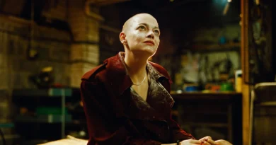 Emma Stone em cena do filme Bugonia, usando jaqueta vermelha em um cenário industrial com tons quentes de ouro e verde. A atriz olha para cima com expressão contemplativa em um ambiente que parece ser uma fábrica ou espaço abandonado. Conteúdo do Multi Nerdz.