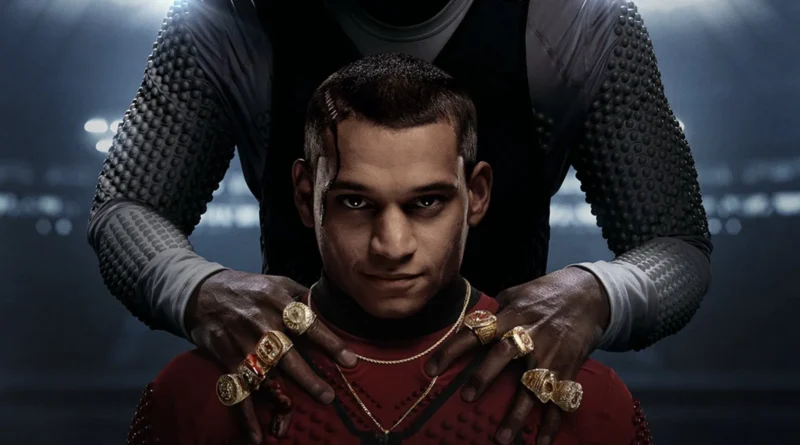 Imagem promocional do filme GOAT (Him) mostra um jovem com expressão séria vestindo uma roupa vermelha, enquanto uma figura maior, com mangas acolchoadas e oito anéis dourados de campeão, segura seus ombros em um ambiente escuro iluminado por luzes de estádio.