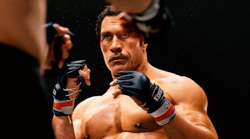 Dwayne “The Rock” Johnson aparece caracterizado como o lutador Mark Kerr em uma cena intensa do filme Coração de Lutador. Com luvas de MMA e expressão séria, ele está em posição de combate dentro do ringue, pronto para enfrentar o oponente. O suor e o sangue em seu rosto destacam a intensidade da luta, enquanto o fundo escuro realça o clima dramático do momento.