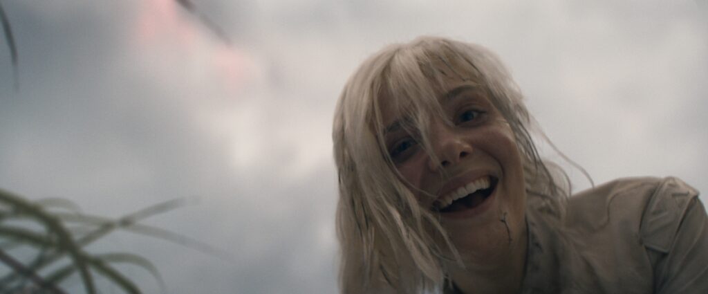 Elle Fanning interpretando a personagem Thia, loira e sorridente, inclinada para frente em meio à névoa cinzenta do céu em cena do filme Predador: Terras Selvagens, com folhagens desfocadas à esquerda.