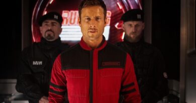 Glen Powell em cena de O Sobrevivente (The Running Man), usando macacão vermelho com o nome Richards no peito em um corredor futurista iluminado de vermelho, escoltado por dois guardas vestidos de preto ao fundo.