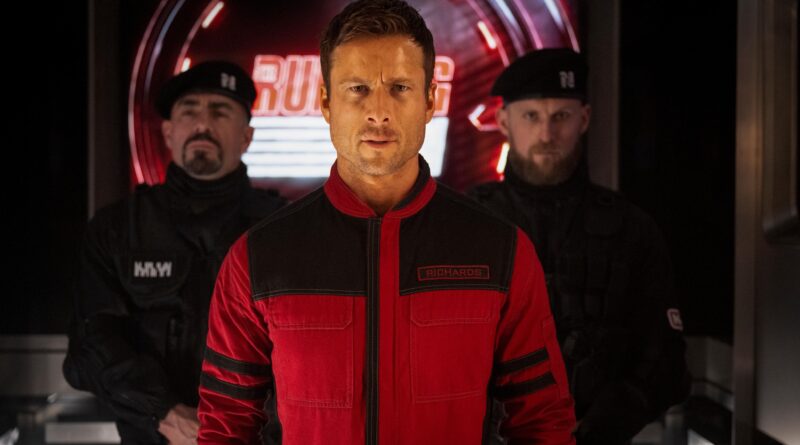 Glen Powell em cena de O Sobrevivente (The Running Man), usando macacão vermelho com o nome Richards no peito em um corredor futurista iluminado de vermelho, escoltado por dois guardas vestidos de preto ao fundo.