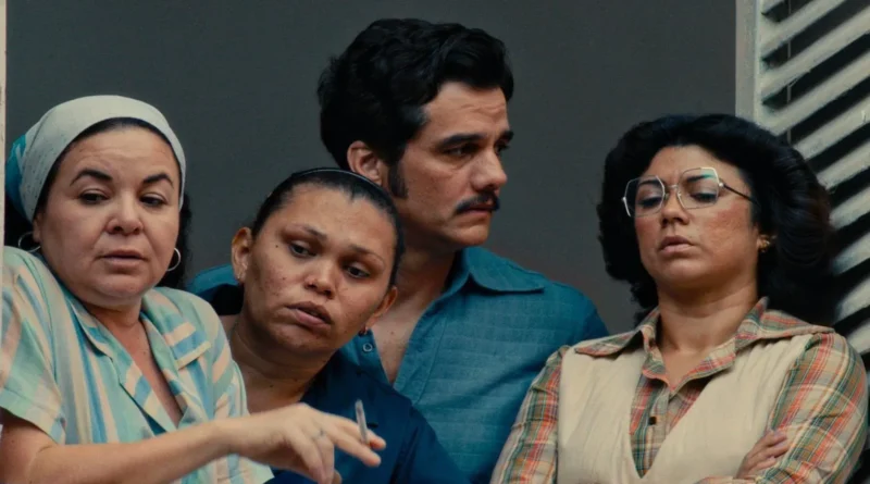 Wagner Moura em cena de O Agente Secreto, de bigode e camisa azul, olhando para o lado enquanto está em uma varanda acompanhado por três mulheres, sendo que uma delas aponta com um cigarro na mão.