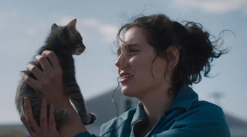 Atriz Eva Victor em cena do filme Sorry Baby, vestindo uma jaqueta azul e com o cabelo preso, segurando um filhote de gato na altura do rosto e olhando para o animal com uma expressão apreensiva ao ar livre.