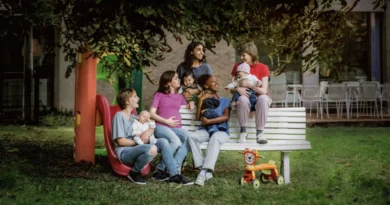 Cena ao ar livre do filme Jovens Mães mostrando cinco jovens mulheres reunidas em torno de um banco branco de praça. Algumas estão grávidas e outras seguram bebês no colo, conversando e sorrindo em um ambiente com árvores, um escorregador colorido e um brinquedo de madeira em forma de leão.