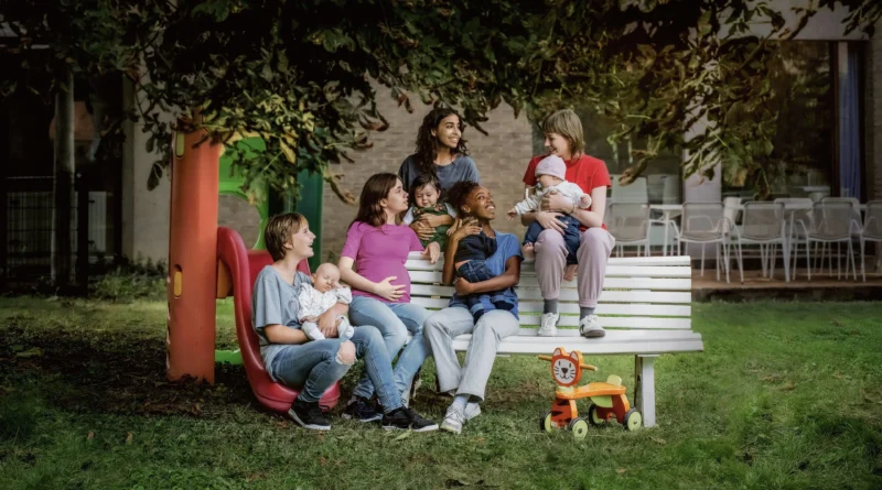 Cena ao ar livre do filme Jovens Mães mostrando cinco jovens mulheres reunidas em torno de um banco branco de praça. Algumas estão grávidas e outras seguram bebês no colo, conversando e sorrindo em um ambiente com árvores, um escorregador colorido e um brinquedo de madeira em forma de leão.