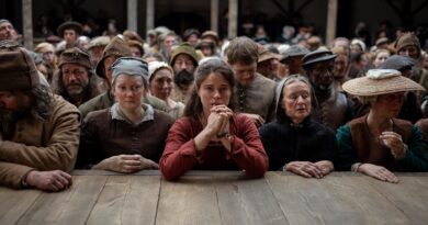 Jessie Buckley em cena ao ar livre do filme Hamnet: A Vida Antes de Hamlet. Ela veste trajes de época rústicos em tons terrosos e está de pé em um ambiente rural, com as mãos unidas à frente do corpo, olhando para o lado com uma expressão pensativa e séria sob luz natural.