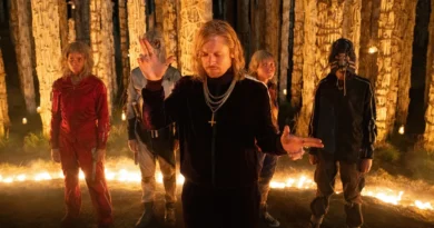 Jack O'Connell como o personagem Jimmy Crystal em Extermínio: Templo dos Ossos, usando um agasalho de veludo escuro e correntes de ouro com um crucifixo. Ele está de braços abertos em um gesto ritualístico, cercado por quatro figuras mascaradas em um ambiente noturno com colunas feitas de ossos e iluminado por velas.