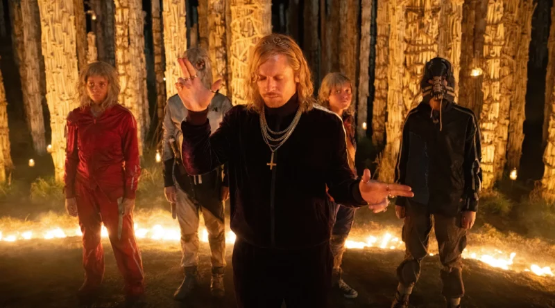 Jack O'Connell como o personagem Jimmy Crystal em Extermínio: Templo dos Ossos, usando um agasalho de veludo escuro e correntes de ouro com um crucifixo. Ele está de braços abertos em um gesto ritualístico, cercado por quatro figuras mascaradas em um ambiente noturno com colunas feitas de ossos e iluminado por velas.