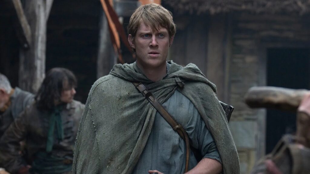 Peter Claffey como Sor Duncan em cena de O Cavaleiro dos Sete Reinos, vestindo uma capa rústica cinza esverdeada com uma alça de couro no peito, olhando para frente com expressão de preocupação em um vilarejo medieval.
