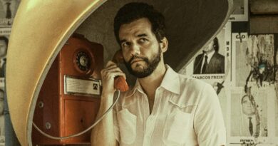 Cena do filme O Agente Secreto mostrando o ator Wagner Moura como o personagem Marcelo. Ele está dentro de um orelhão público amarelo e laranja clássico, segurando o monofone preto na orelha direita enquanto fala. Ele aparece de bigode espesso e veste uma jaqueta de veludo cotelê marrom sobre uma camisa xadrez escura, olhando para a esquerda com uma expressão tensa, observadora e preocupada. A rua brasileira dos anos 70 ao fundo está movimentada, com carros antigos (Fusca, Opala), pessoas e cartazes de rua visíveis mas desfocados (bokeh). A fotografia tem granulação de filme vintage e cores quentes.