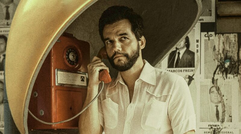 Cena do filme O Agente Secreto mostrando o ator Wagner Moura como o personagem Marcelo. Ele está dentro de um orelhão público amarelo e laranja clássico, segurando o monofone preto na orelha direita enquanto fala. Ele aparece de bigode espesso e veste uma jaqueta de veludo cotelê marrom sobre uma camisa xadrez escura, olhando para a esquerda com uma expressão tensa, observadora e preocupada. A rua brasileira dos anos 70 ao fundo está movimentada, com carros antigos (Fusca, Opala), pessoas e cartazes de rua visíveis mas desfocados (bokeh). A fotografia tem granulação de filme vintage e cores quentes.