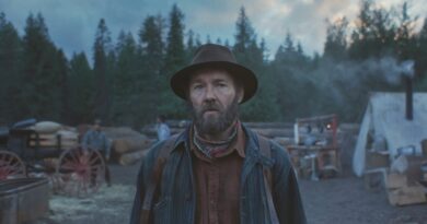 Joel Edgerton caracterizado como o protagonista Robert Grainier em cena de Sonhos de Trem (Train Dreams), para a capa da crítica do blog Multi Nerdz. Ele está de pé ao crepúsculo em um acampamento florestal, olhando para a câmera com expressão séria. Ele usa um chapéu de feltro marrom, jaqueta listrada e bandana. No fundo desfocado, há uma carroça e uma barraca de lona branca entre árvores perenes.