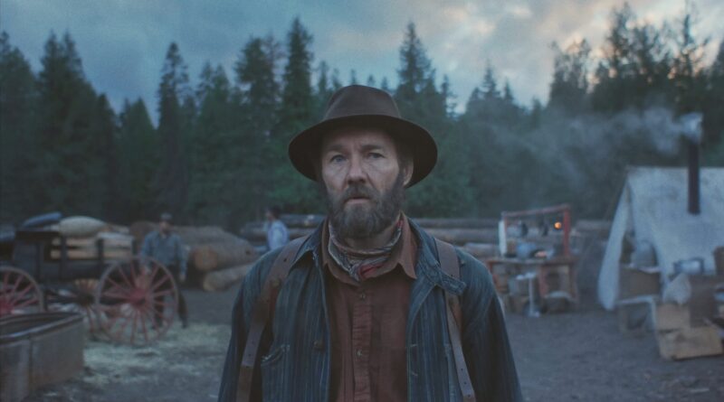 Joel Edgerton caracterizado como o protagonista Robert Grainier em cena de Sonhos de Trem (Train Dreams), para a capa da crítica do blog Multi Nerdz. Ele está de pé ao crepúsculo em um acampamento florestal, olhando para a câmera com expressão séria. Ele usa um chapéu de feltro marrom, jaqueta listrada e bandana. No fundo desfocado, há uma carroça e uma barraca de lona branca entre árvores perenes.