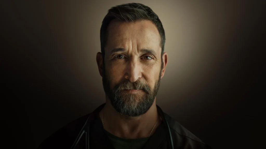 Retrato em médio-primeiro plano do ator Noah Wyle, protagonista da série 'The Pitt'. Ele tem uma barba grisalha e marrom cheia, olhos castanhos expressivos e uma expressão séria olhando diretamente para a câmera. Ele veste um uniforme médico verde-escuro e um estetoscópio está pendurado em seu pescoço. Uma fina corrente de ouro com um pingente é visível sob a gola. O fundo é um gradiente escuro e suave, com iluminação central focada em seu rosto. Capa da crítica de 'The Pitt' no blog Multi Nerdz.