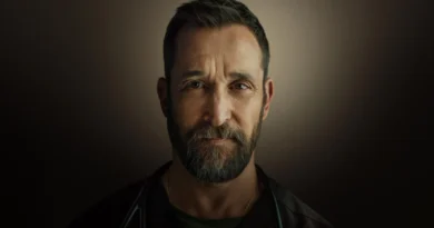 Retrato em médio-primeiro plano do ator Noah Wyle, protagonista da série 'The Pitt'. Ele tem uma barba grisalha e marrom cheia, olhos castanhos expressivos e uma expressão séria olhando diretamente para a câmera. Ele veste um uniforme médico verde-escuro e um estetoscópio está pendurado em seu pescoço. Uma fina corrente de ouro com um pingente é visível sob a gola. O fundo é um gradiente escuro e suave, com iluminação central focada em seu rosto. Capa da crítica de 'The Pitt' no blog Multi Nerdz.