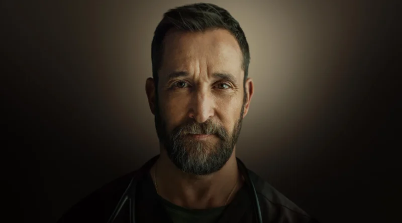Retrato em médio-primeiro plano do ator Noah Wyle, protagonista da série 'The Pitt'. Ele tem uma barba grisalha e marrom cheia, olhos castanhos expressivos e uma expressão séria olhando diretamente para a câmera. Ele veste um uniforme médico verde-escuro e um estetoscópio está pendurado em seu pescoço. Uma fina corrente de ouro com um pingente é visível sob a gola. O fundo é um gradiente escuro e suave, com iluminação central focada em seu rosto. Capa da crítica de 'The Pitt' no blog Multi Nerdz.