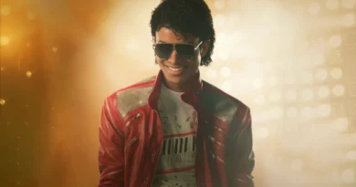 Foto de Jaafar Jackson caracterizado como Michael Jackson no filme cinebiográfico 'Michael'. Ele está sorrindo, usando óculos de sol aviador e a icônica jaqueta de couro vermelha com ombreiras prateadas texturizadas. Por baixo, uma camiseta branca com teclas de piano e notas musicais impressas. Ele está em um palco, com fortes luzes douradas e nebulosas ao fundo, criando uma atmosfera de show. Capa da crítica do filme no blog Multi Nerdz.