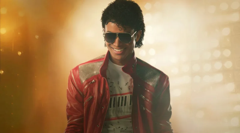 Foto de Jaafar Jackson caracterizado como Michael Jackson no filme cinebiográfico 'Michael'. Ele está sorrindo, usando óculos de sol aviador e a icônica jaqueta de couro vermelha com ombreiras prateadas texturizadas. Por baixo, uma camiseta branca com teclas de piano e notas musicais impressas. Ele está em um palco, com fortes luzes douradas e nebulosas ao fundo, criando uma atmosfera de show. Capa da crítica do filme no blog Multi Nerdz.