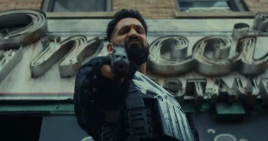 Imagem em contrapicado (ângulo de baixo para cima) do ator Jon Bernthal como Frank Castle no filme 'O Justiceiro: Uma Última Morte'. Ele tem uma expressão dura, intimidadora e com o cenho franzido, apontando uma pistola preta diretamente para a câmera. Ele veste um equipamento tático preto, incluindo luvas e um colete com bolsos de munição, onde parte do seu icônico símbolo da caveira branca está pintado de forma rústica. Ao fundo, levemente desfocado, há a fachada de um prédio de tijolos com um grande letreiro antigo em metal e neon. Capa da crítica do filme no blog Multi Nerdz.