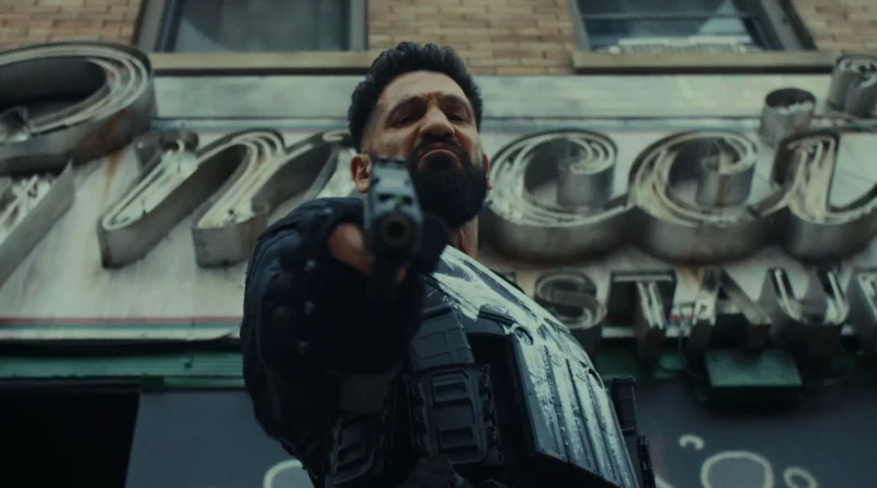 Imagem em contrapicado (ângulo de baixo para cima) do ator Jon Bernthal como Frank Castle no filme 'O Justiceiro: Uma Última Morte'. Ele tem uma expressão dura, intimidadora e com o cenho franzido, apontando uma pistola preta diretamente para a câmera. Ele veste um equipamento tático preto, incluindo luvas e um colete com bolsos de munição, onde parte do seu icônico símbolo da caveira branca está pintado de forma rústica. Ao fundo, levemente desfocado, há a fachada de um prédio de tijolos com um grande letreiro antigo em metal e neon. Capa da crítica do filme no blog Multi Nerdz.