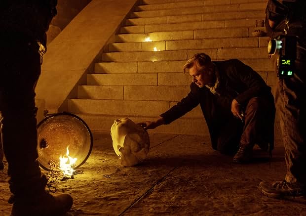 Christopher Nolan durante as gravações de ‘A Odisseia’, agachado ao lado de uma caveira cenográfica e de uma estrutura metálica em chamas, com escadarias e integrantes da equipe ao redor.