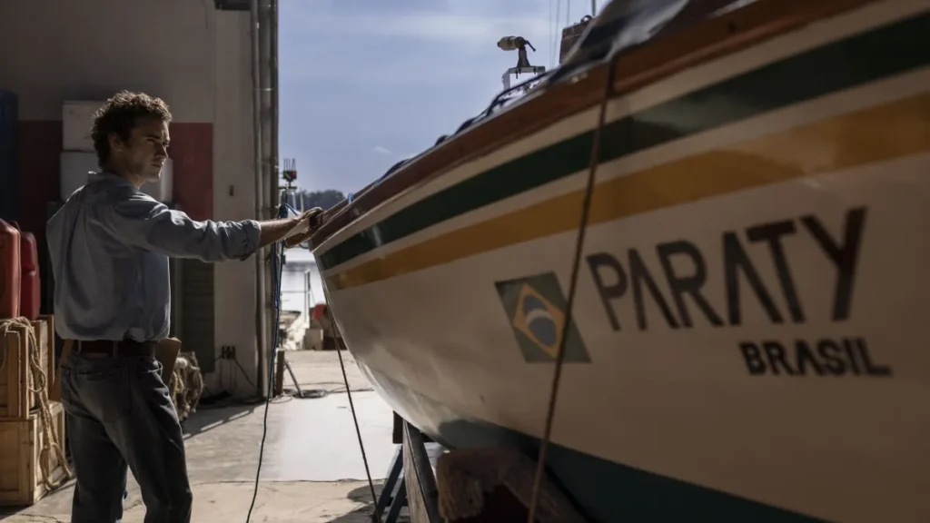 Filipe Bragança como Amyr Klink aparece ao lado do barco Paraty Brasil, tocando a embarcação em um galpão iluminado pela luz do dia, em imagem do filme ‘100 Dias’.