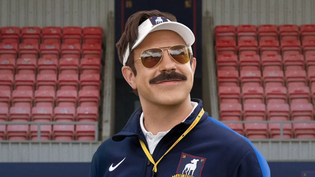 Jason Sudeikis caracterizado como Ted Lasso aparece sorrindo em um estádio, usando óculos escuros, viseira branca e agasalho esportivo azul-marinho do AFC Richmond.