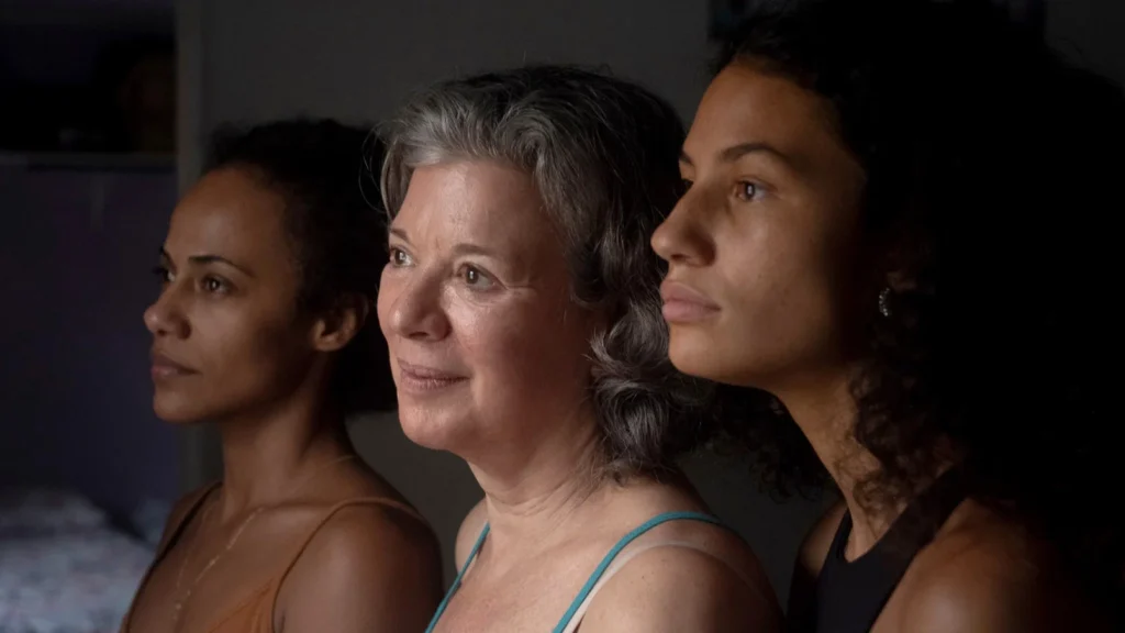 Carla Ribas como Dolores, Naruna Costa como Deborah e Ariane Aparecida como Duda aparecem juntas em uma cena intimista do filme ‘Dolores’, representando três gerações da mesma família enquanto observam algo fora de cena.