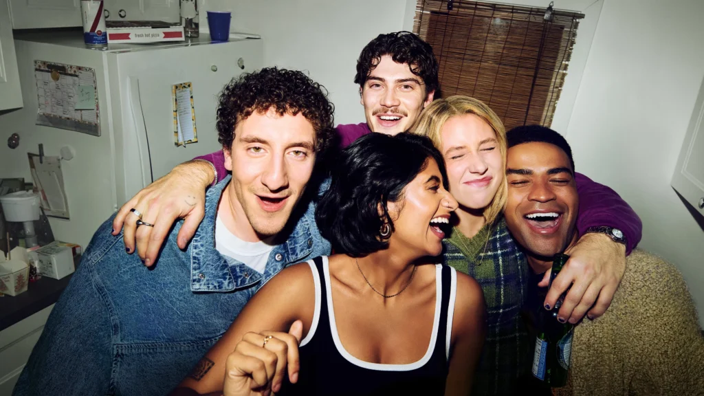 Os protagonistas da série ‘Adults’ aparecem abraçados e sorrindo para a câmera em uma foto descontraída tirada em uma cozinha, com clima de amizade e celebração.