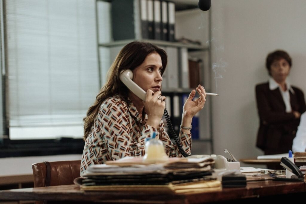 Mel Lisboa como a jornalista Silvana aparece sentada em um escritório, falando ao telefone enquanto segura um cigarro aceso, em cena do filme ‘A Conspiração Condor’.