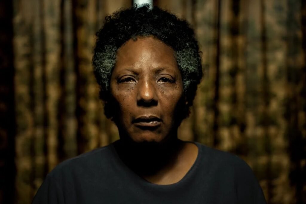 Rejane Faria como Vicentina aparece em primeiro plano, encarando a câmera com expressão séria, diante de um fundo escuro e desfocado, em imagem do filme ‘Vicentina Pede Desculpas’.
