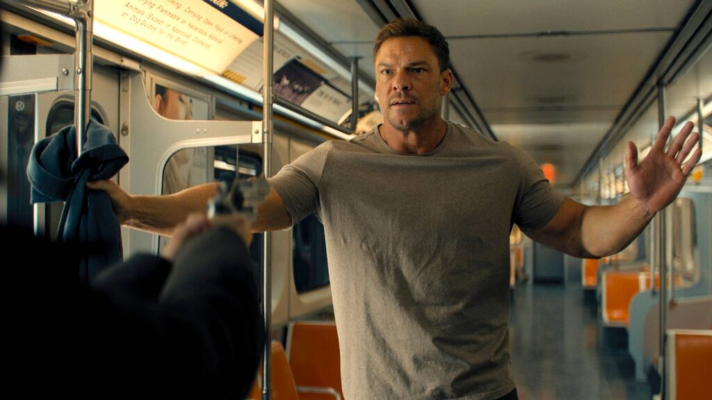 Alan Ritchson como Jack Reacher aparece dentro de um vagão de metrô, com os braços abertos e expressão tensa, enquanto uma arma é apontada em sua direção em cena da série ‘Reacher’.