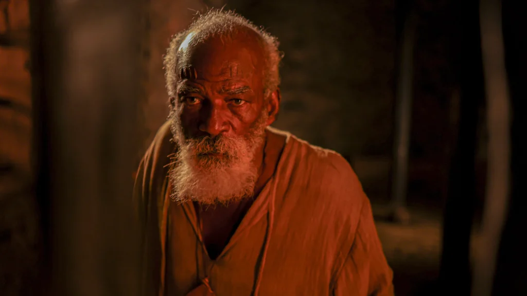 Antonio Pitanga como Pacífico Licutan aparece em destaque com barba branca, marcas no rosto e expressão séria, iluminado por tons quentes e avermelhados, em cena do filme ‘Malês’.