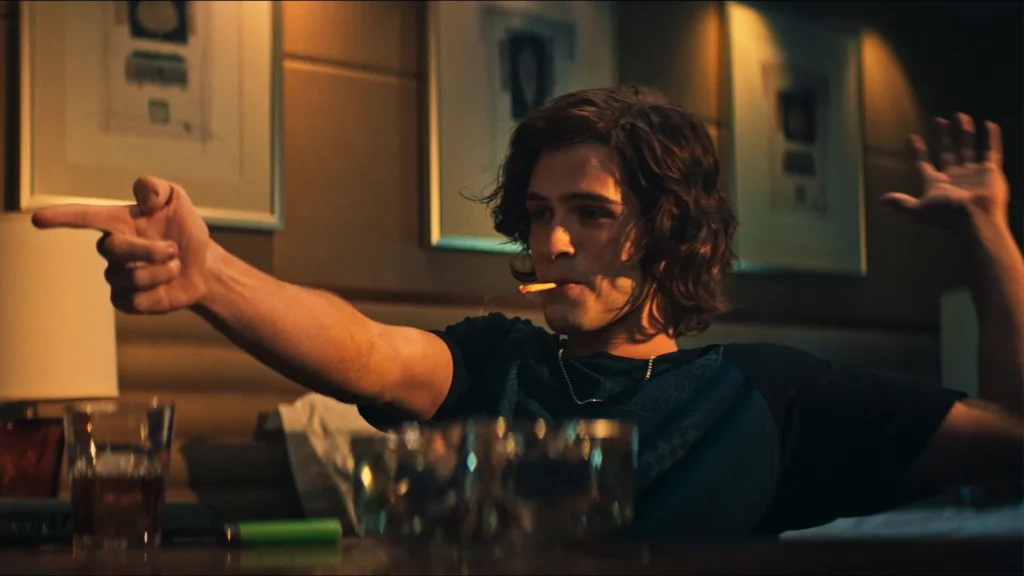 João Guilherme Ávila como Daniel Nascimento aparece sentado em um ambiente com luz baixa, fazendo um gesto com a mão em forma de arma e segurando um cigarro na boca, em imagem do filme ‘O Rei da Internet’.