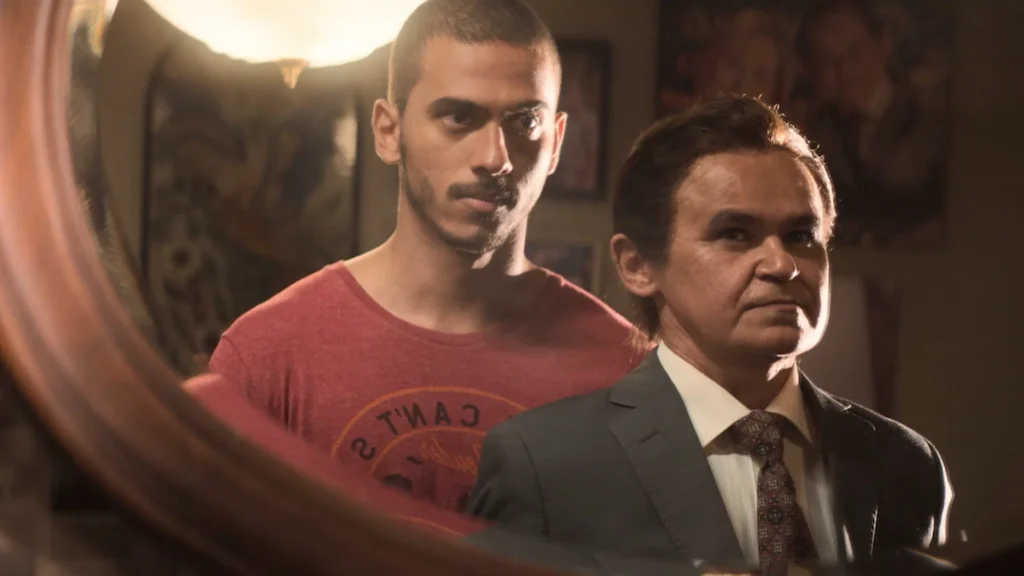Cena do filme ‘Papagaios’ mostra dois homens refletidos em um espelho, um usando camiseta vermelha e outro de terno e gravata, em um ambiente iluminado por luz quente. O longa é protagonizado por Gero Camilo como Tunico e Ruan Aguiar como Beto.